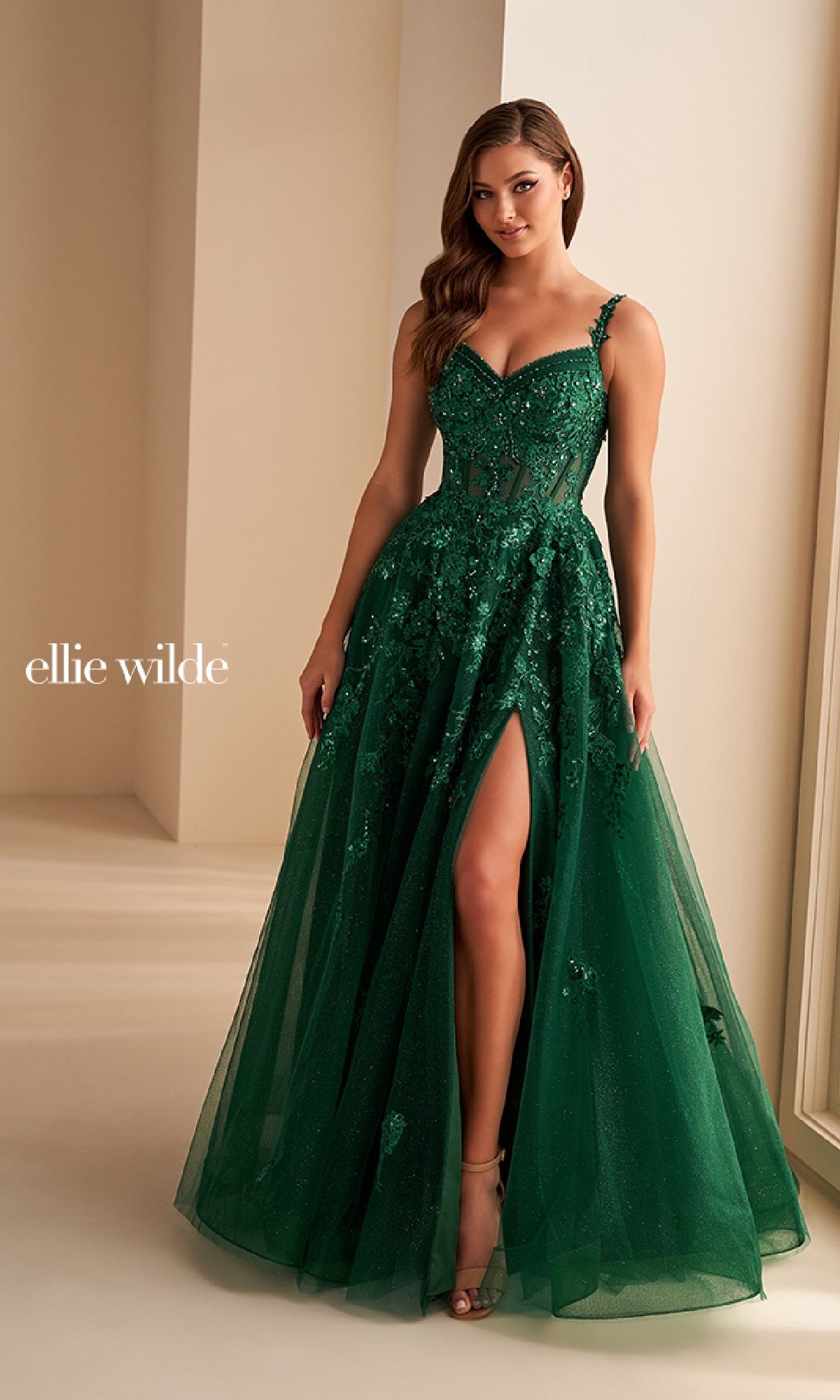 A woman stands indoors near large windows wearing the Ellie Wilde EW36204, a deep green long prom dress featuring embellishments, a sleeveless design, thigh-high slit, sheer panels, embroidery, and delicate floral lace appliques.